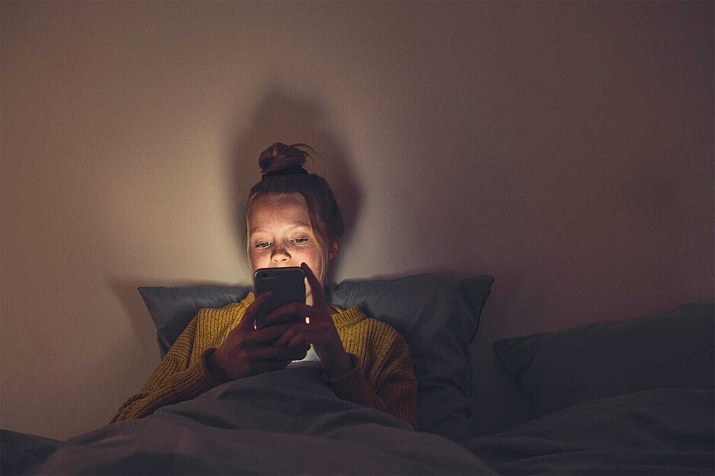 Das erste Smartphone für Ihr Kind – Tipps für Eltern Mädchen im Dunkeln auf Bett, schaut auf Smartphone-Display
