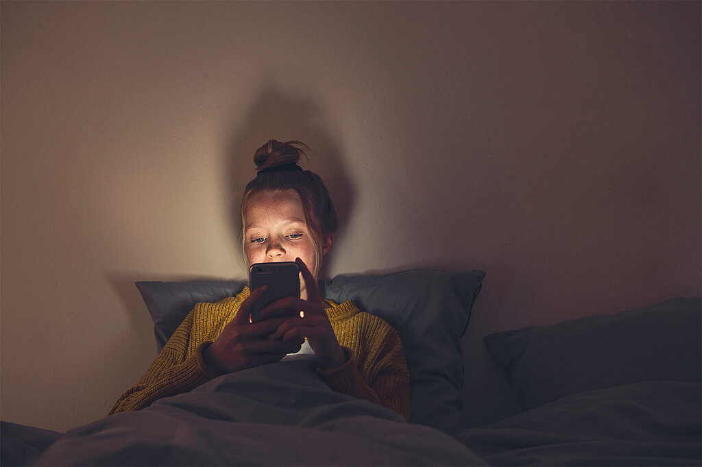 Mädchen im Dunkeln auf Bett, schaut auf Smartphone-Display
