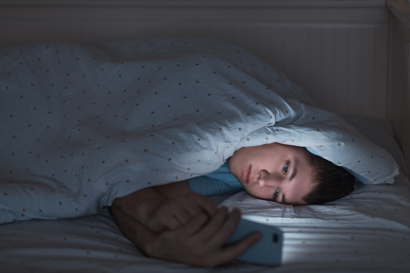 Junge unter Bettdecke schaut müde auf Smartphone