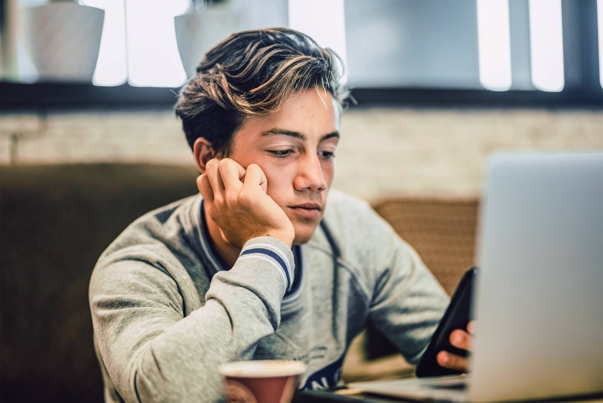 Junge vor Laptop schaut deprimiert auf Smartphone